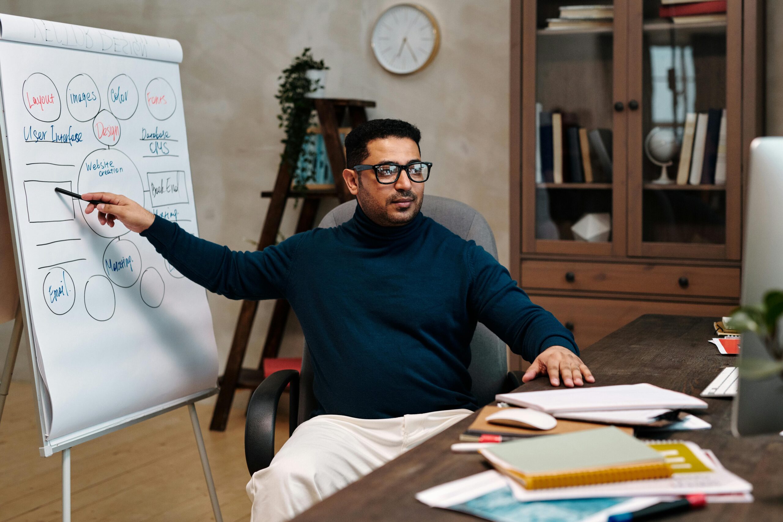 Man teaching online, pointing at a flipchart in a cozy office space. Perfect for online education themes.